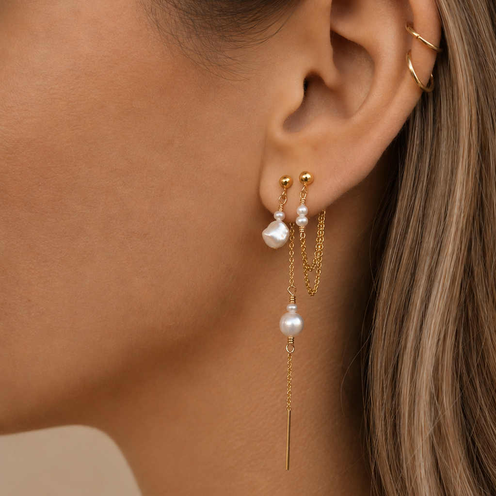 A close-up of a model's ear wearing a pair of gold drop earrings with white freshwater pearls, along with a gold hoop earring in the opposite ear.
