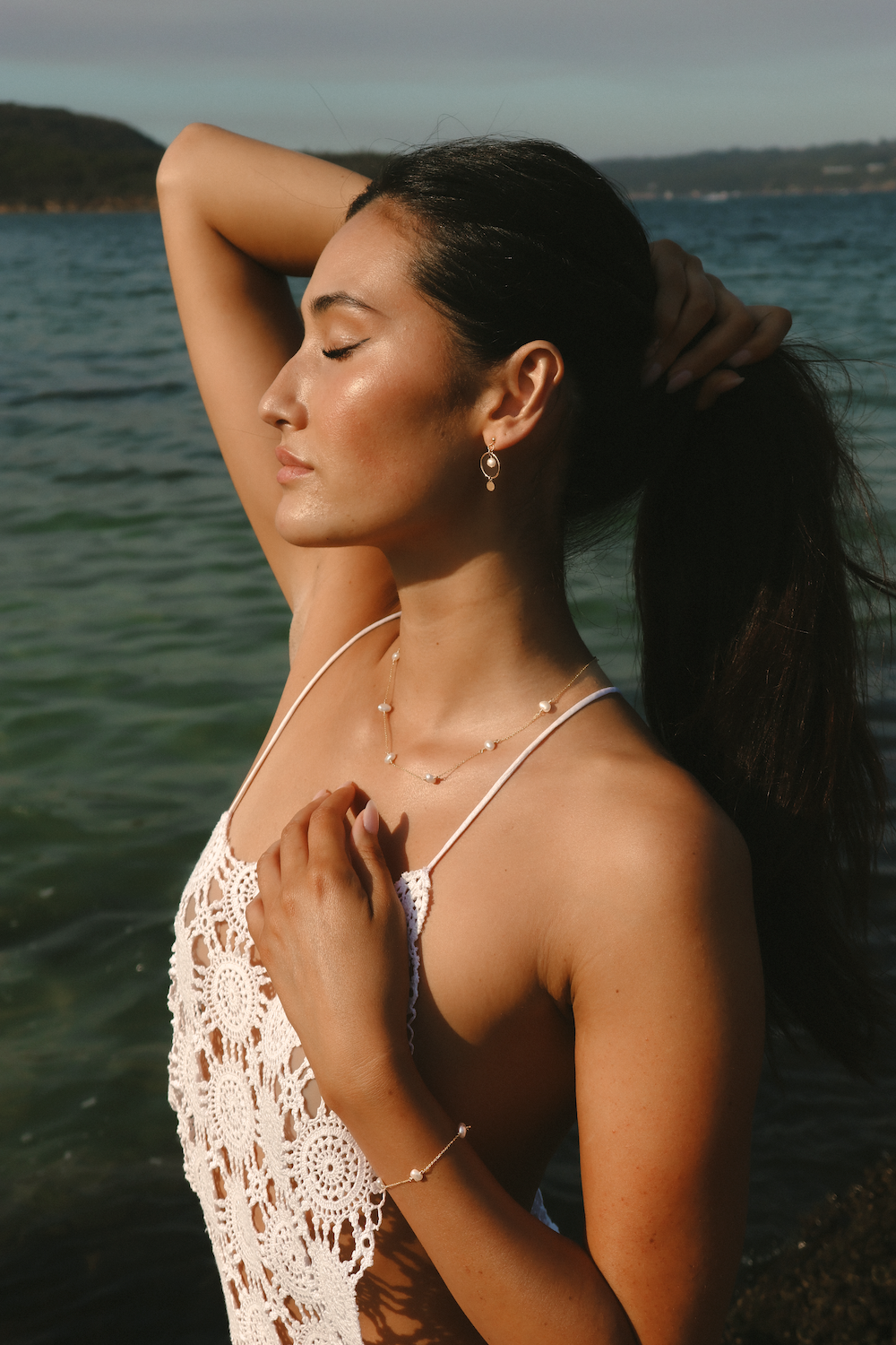 A woman with her hair in a ponytail stands by the water wearing delicate pearl necklaces, a pearl bracelet, and a white crochet halter top.