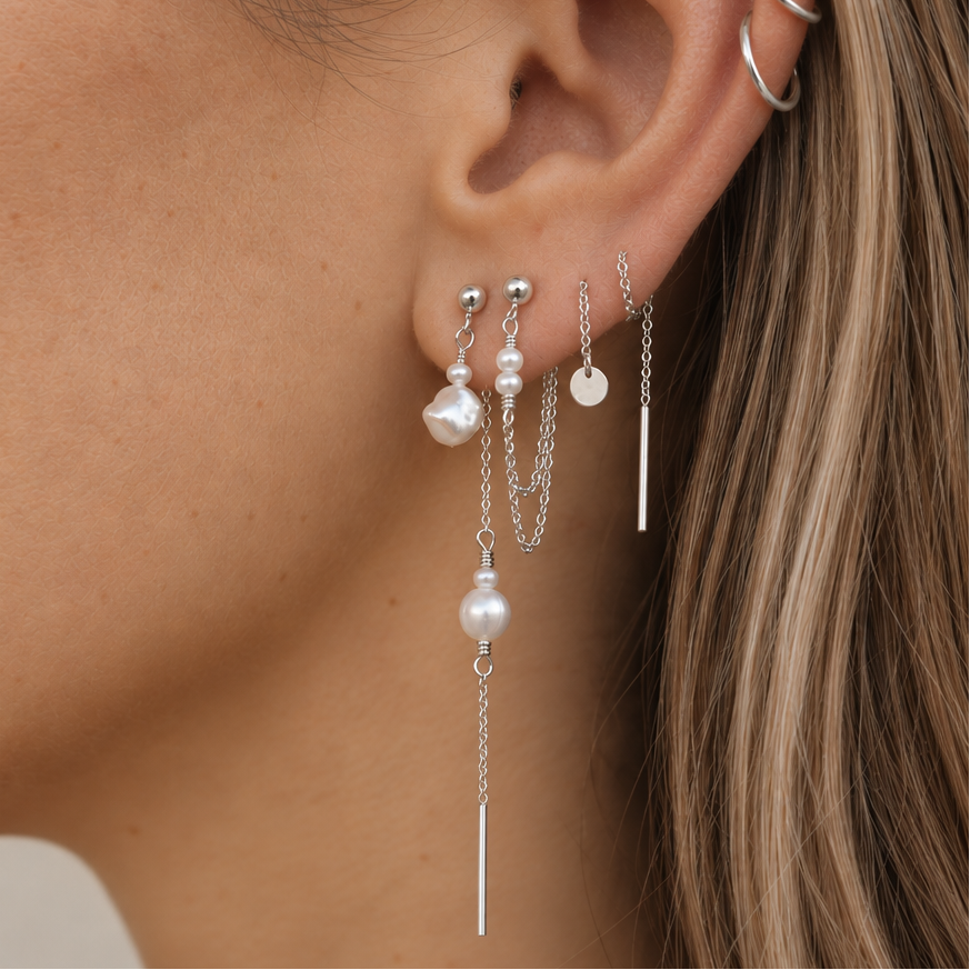 A close-up of a model's ear wearing a multi-piece silver ear cuff and layered dangle earrings with white pearl beads and silver chains.
