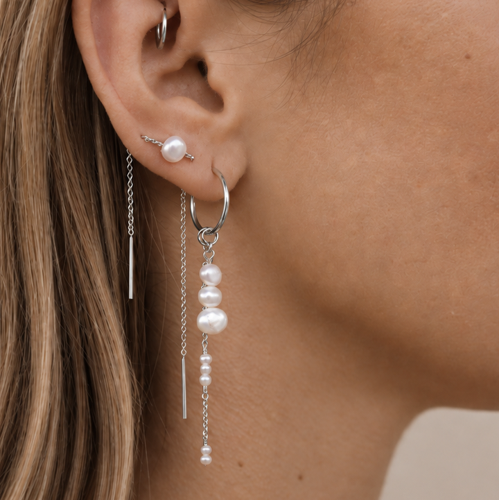 A close-up side profile of a woman's ear wearing a silver hoop earring with dangling white pearl beads and a small silver stud earring.
