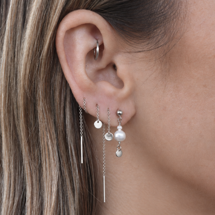 A close-up of a woman's ear wearing a pair of silver dangle earrings with small pearl-like beads and thin chain drops.